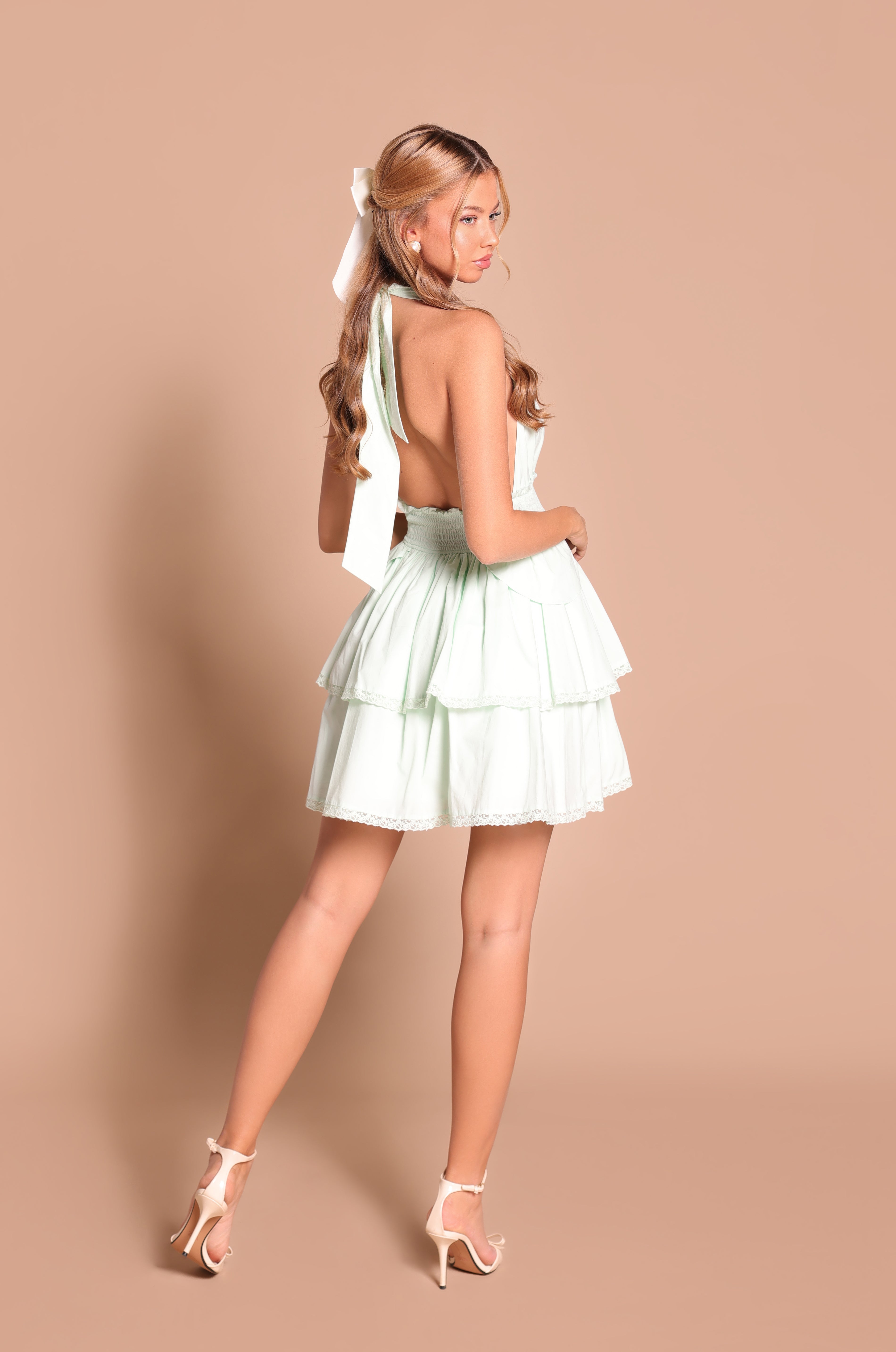 Woman wearing the Serenya Mint Vista Pastel Halter Open-Back Sundress with a large white bow in her hair and heels, standing against a beige background facing away from the camera.